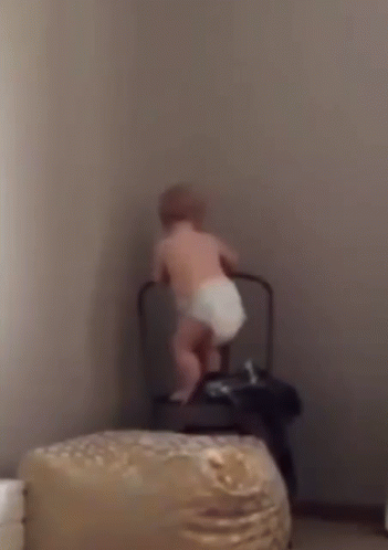 A Baby In A Diaper Is Standing Next To A Chair . GIF