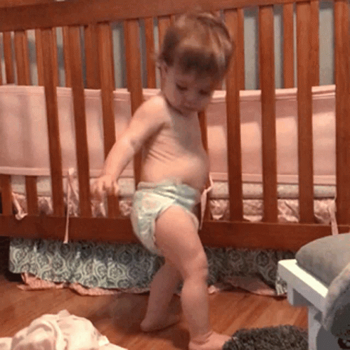 A Baby In A Diaper Is Standing Next To A Crib GIF