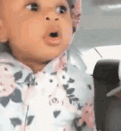 A Baby In A Floral Outfit Is Sitting In A Car Seat With Her Mouth Open . GIF