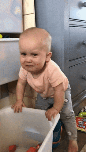 A Baby In A Pink Shirt Is Standing Next To A White Bin With Toys In It GIF