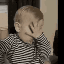 A Baby In A Striped Shirt Is Covering His Face With His Hands . GIF