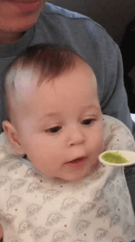 A Baby Is Being Fed With A Spoon By A Man In A Blue Shirt GIF