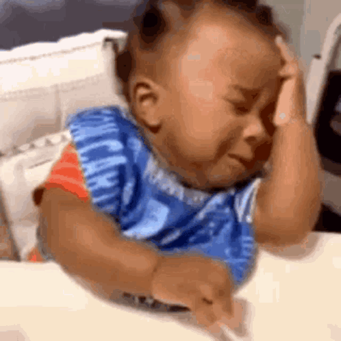 A Baby Is Crying While Sitting At A Table With His Hands On His Head . GIF