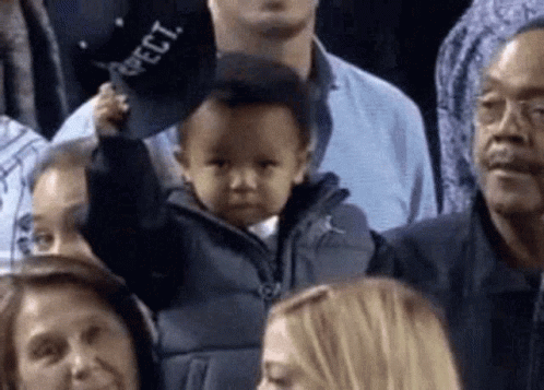 A Baby Is Holding A Hat In A Crowd Of People At A Basketball Game . GIF
