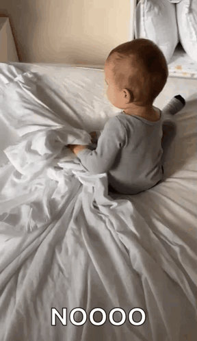 A Baby Is Laying On A Bed With A White Blanket And The Word Nooo Written On It GIF