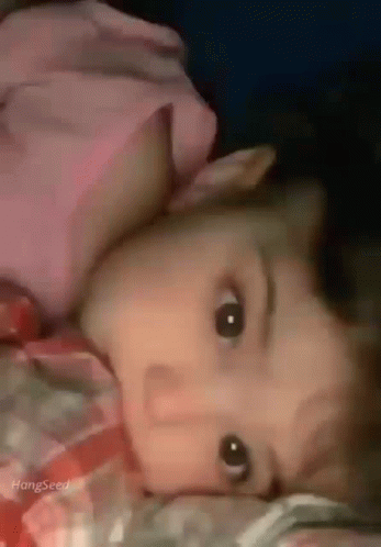 A Baby Is Laying On A Bed With His Head On A Pillow And Looking At The Camera . GIF