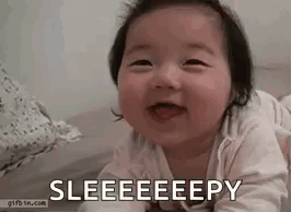 A Baby Is Laying On Its Stomach On A Bed And Smiling With Its Tongue Out . GIF