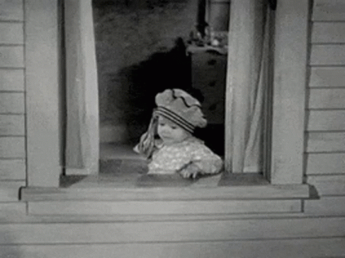 A Baby Is Looking Out Of A Window Wearing A Hat . GIF
