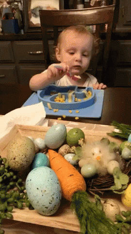 A Baby Is Sitting At A Table With A Plate Of Food And A Spoon GIF