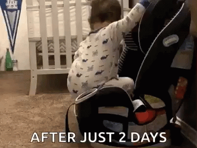 A Baby Is Sitting In A Car Seat Next To A Crib In A Bedroom . GIF