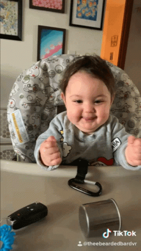 A Baby Is Sitting In A High Chair With A Sticker On His Shirt That Says Anna GIF