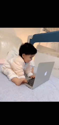 A Baby Is Sitting On A Bed Using An Apple Laptop Computer . GIF