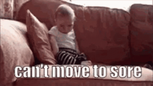 A Baby Is Sitting On A Couch With The Words `` Can 'T Move To Sore '' Written On The Bottom . GIF
