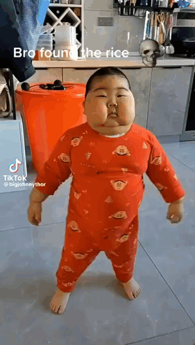 A Baby Is Standing In A Kitchen Wearing A Red Pajama Set . GIF
