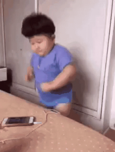 A Baby Is Standing On A Bed Next To A Cell Phone . GIF