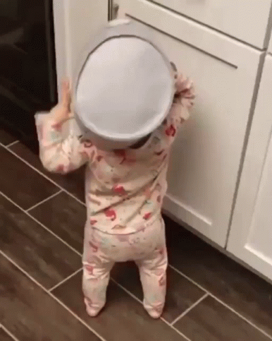 A Baby Is Standing On A Tiled Floor Holding A Pot On Her Head . GIF