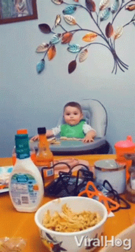A Baby Sits In A High Chair Next To A Bowl Of Noodles GIF
