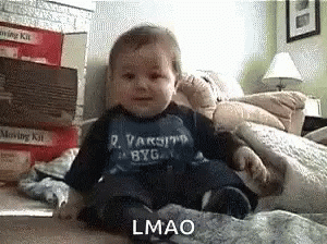 A Baby Wearing A Blue Varsity Byg Shirt Is Sitting On A Bed . GIF