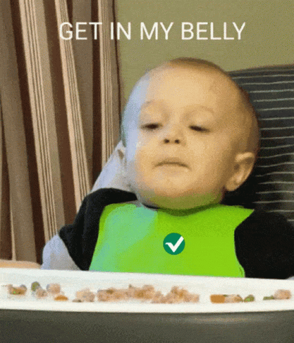 A Baby Wearing A Green Bib Is Sitting At A Table With A Plate Of Food And The Words Get In My Belly GIF