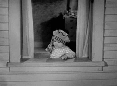 A Baby Wearing A Hat Looks Out A Window GIF