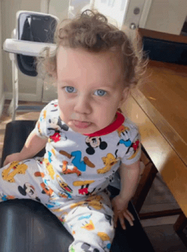 A Baby Wearing A Mickey Mouse Pajama Is Sitting On A Chair GIF