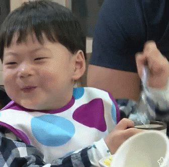 A Baby Wearing A Polka Dot Bib Is Smiling While Eating From A Bowl . GIF