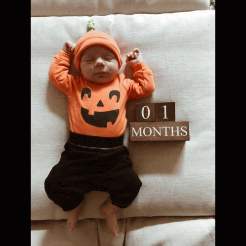 A Baby Wearing A Pumpkin Outfit Is Sleeping Next To A Sign That Says 1 Months GIF