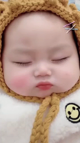 A Baby Wearing A Yellow Knitted Hat And A White Bib With A Smiley Face On It . GIF