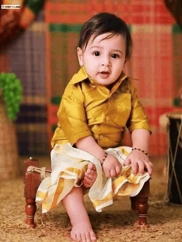 A Baby Wearing A Yellow Shirt And White Skirt Is Sitting On A Small Wooden Chair GIF