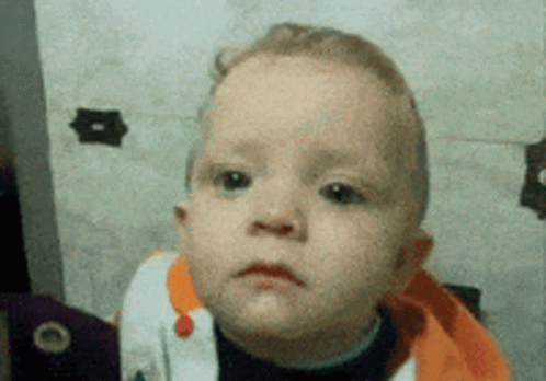 A Baby With A Sad Look On His Face Is Wearing An Orange Sweater GIF