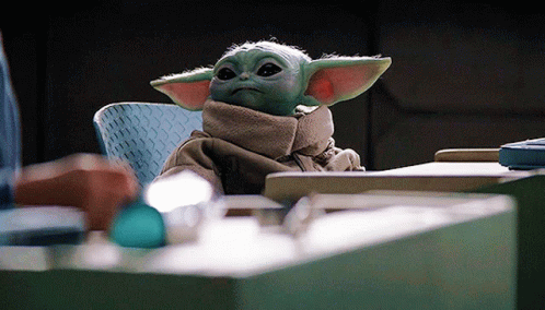 A Baby Yoda Is Sitting At A Table With A Scarf Around His Neck . GIF
