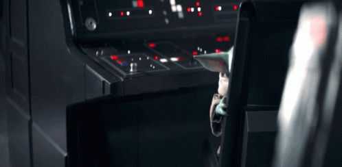 A Baby Yoda Is Sitting In A Chair In Front Of A Control Panel In A Dark Room . GIF