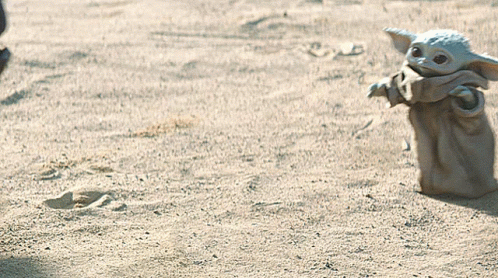 A Baby Yoda Toy Is Standing In The Sand Looking At Something GIF