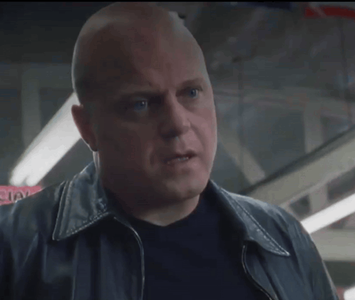 A Bald Man In A Black Jacket Looks Up At Something GIF