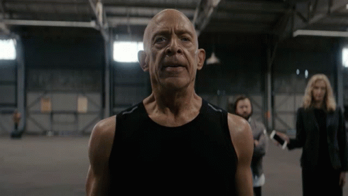 A Bald Man In A Black Tank Top Is Standing In A Dark Room GIF
