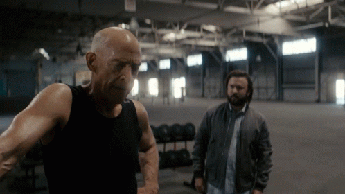 A Bald Man In A Black Tank Top Stands Next To Another Man In A Grey Jacket GIF