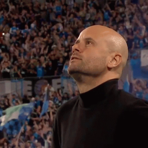 A Bald Man In A Black Turtleneck Stands In Front Of A Crowd And Looks Up GIF