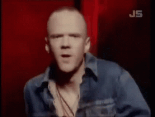 A Bald Man In A Blue Denim Jacket Is Standing In Front Of A Red Background . GIF