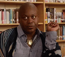 A Bald Man In A Blue Shirt And Striped Sweater Is Standing In Front Of A Bookshelf . GIF