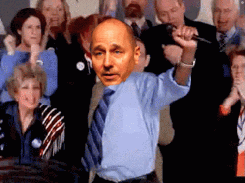 A Bald Man In A Blue Shirt And Tie Holds A Microphone In Front Of A Crowd GIF