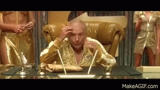 A Bald Man In A Gold Robe Is Sitting At A Desk With A Pen In His Hand . GIF