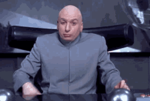 A Bald Man In A Gray Suit Is Sitting In A Chair And Making A Face . GIF
