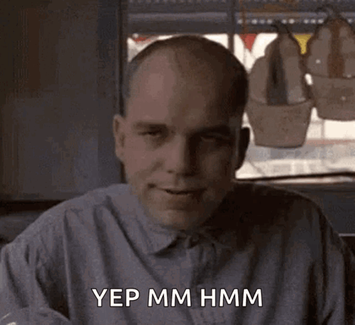 A Bald Man In A Grey Shirt Is Sitting In Front Of A Window And Saying Yep Mm Hmm GIF