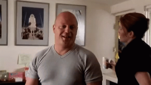 A Bald Man In A Grey Shirt Is Smiling While A Woman Stands Behind Him GIF