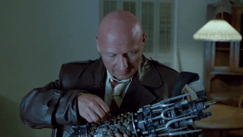 A Bald Man In A Leather Jacket And Tie Is Holding A Mechanical Arm GIF