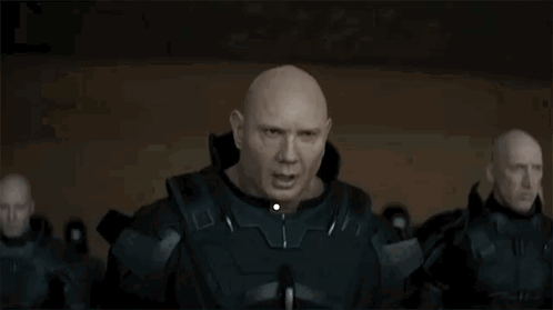 A Bald Man In A Military Uniform Is Standing In Front Of A Group Of Bald Men In A Dark Room . GIF