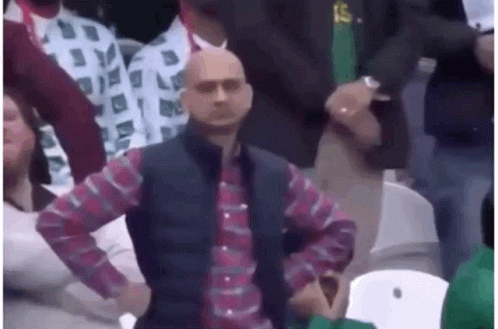 A Bald Man In A Plaid Shirt And Vest Is Sitting In A Crowd . GIF