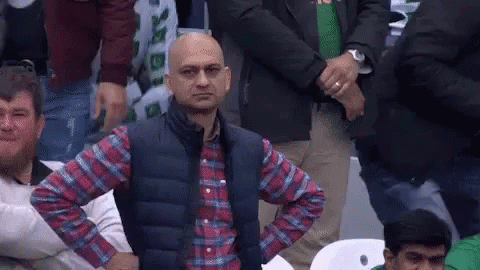 A Bald Man In A Plaid Shirt And Vest Is Sitting In A Crowd With His Hands On His Hips . GIF