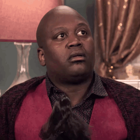 A Bald Man In A Red Vest Is Sitting In Front Of A Lamp And Making A Funny Face . GIF