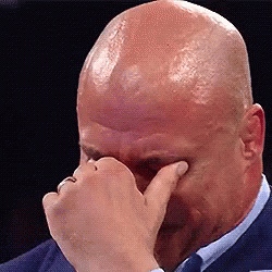 A Bald Man In A Suit And Tie Is Covering His Eyes With His Hand . GIF